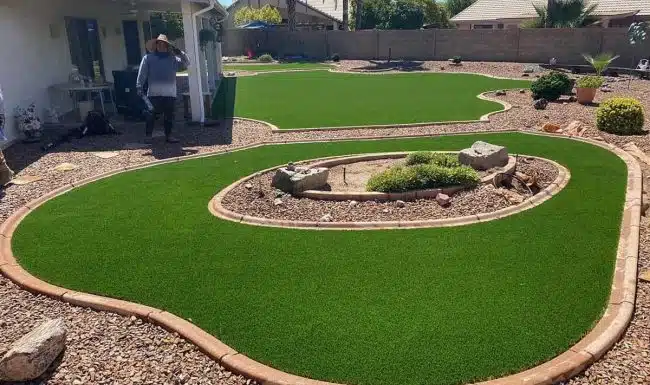 Large residential backyard with expansive artificial turf lawn and a curved, raised rock garden flower bed.